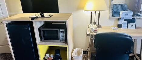 Habitación de hotel con una cama, un escritorio con lámpara, microondas, cafetera y un cubo de basura.