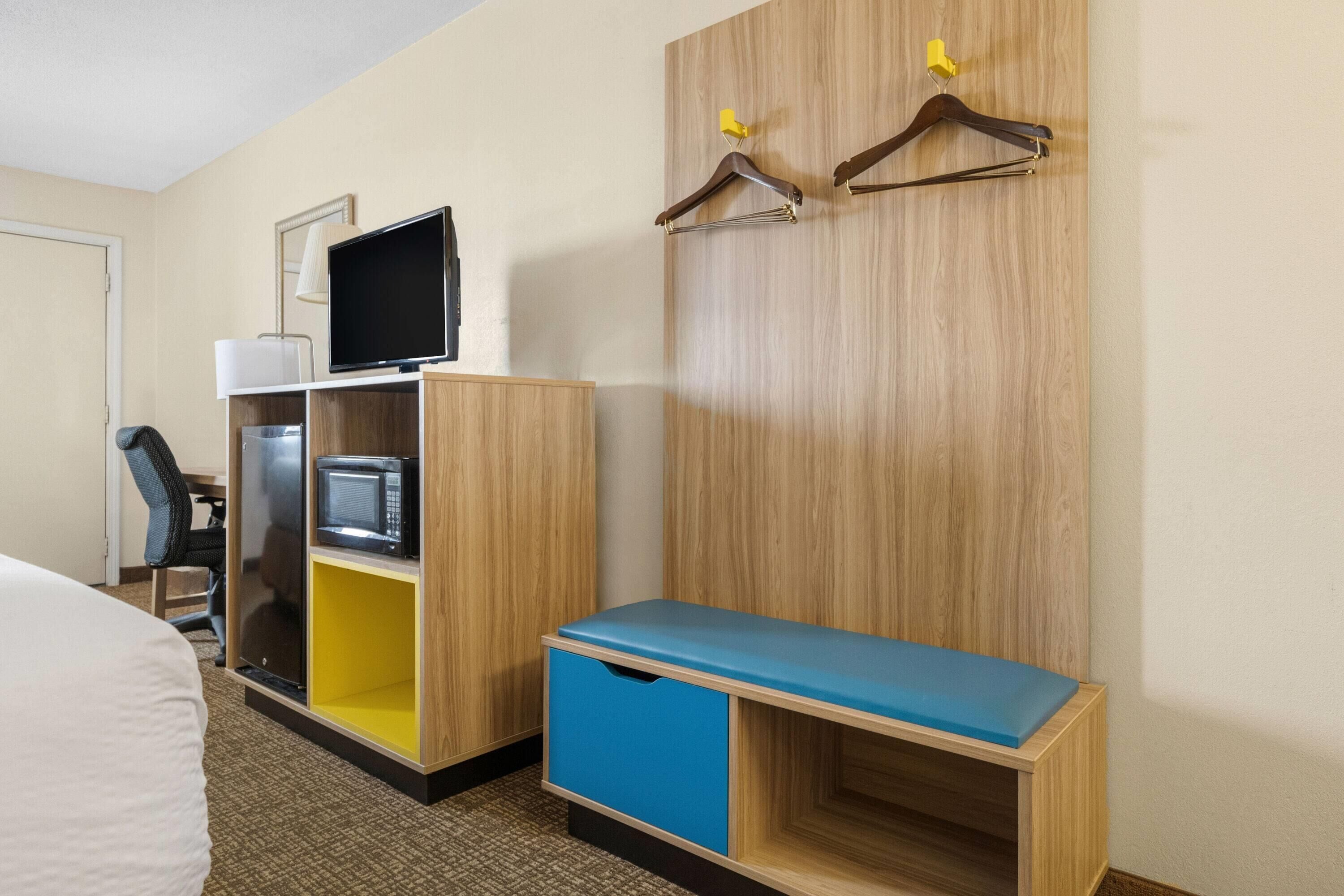 A hotel room with a bed, a desk, a chair, a wardrobe with a TV and microwave, and a bench.