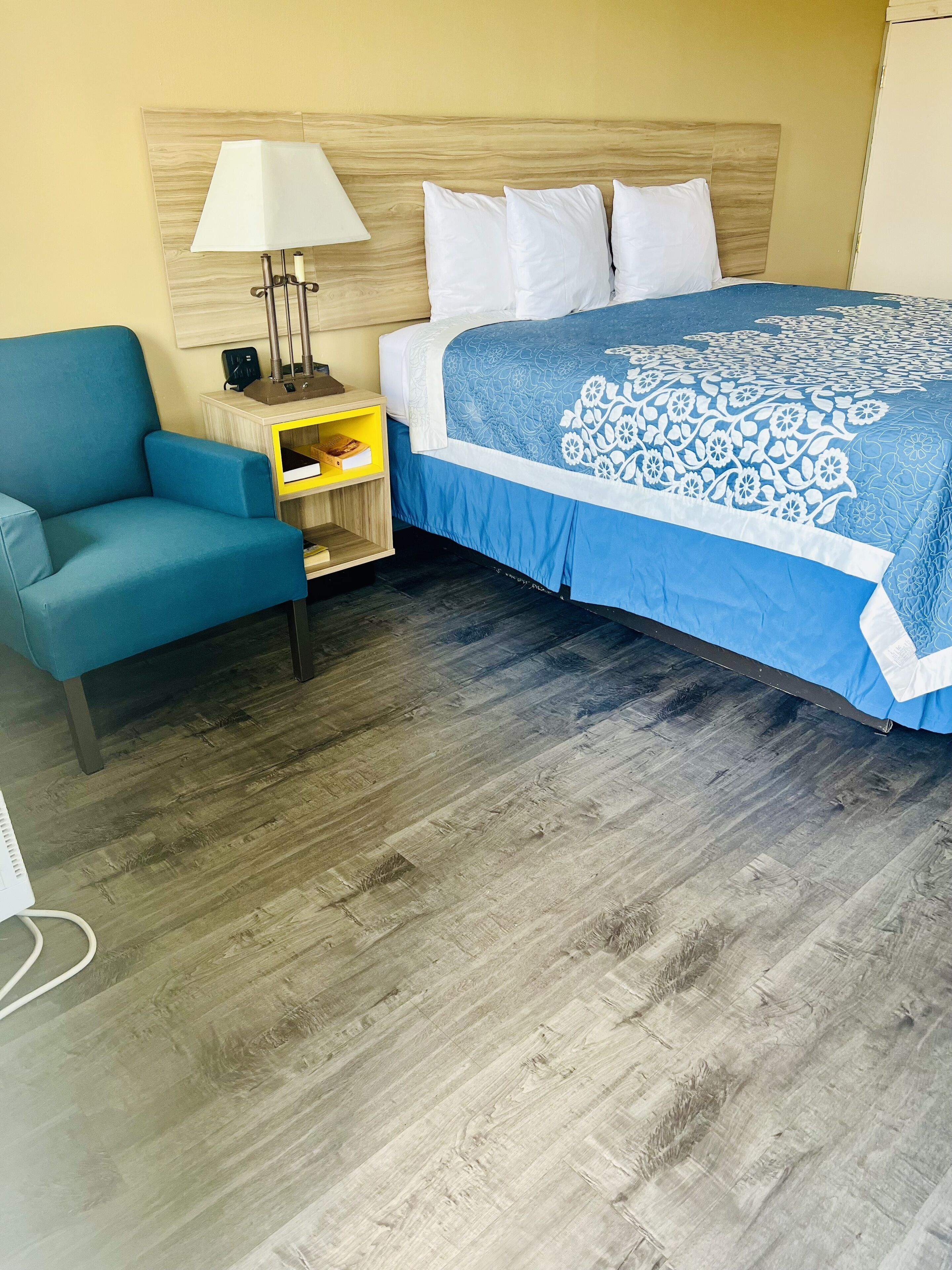 Una habitación de hotel con una cama, un sillón azul, una mesita de noche con una lámpara y un cabecero de madera.