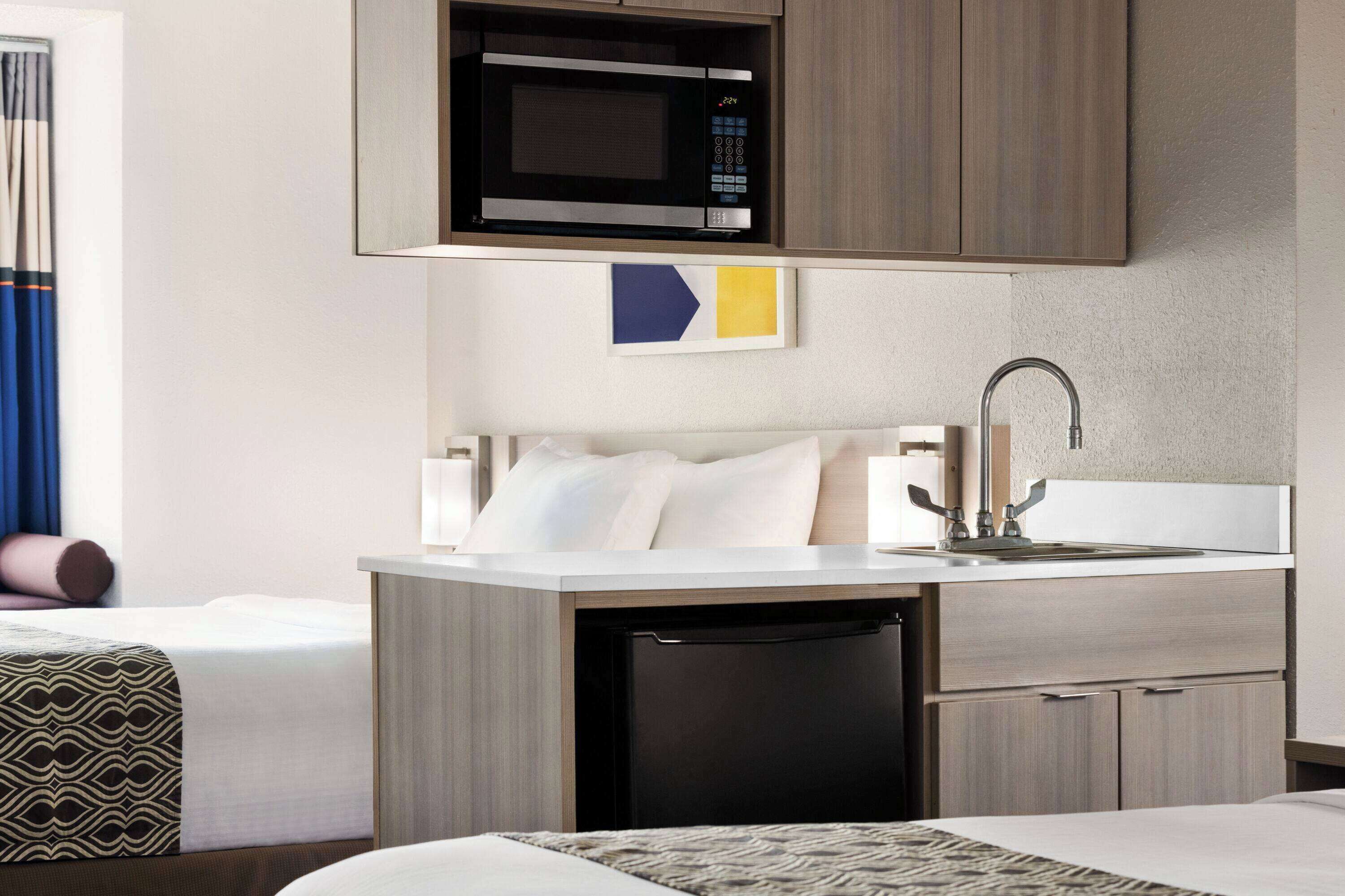 Habitación de hotel compacta con kitchenette, que incluye microondas, lavaplatos y lavabo. Se observa una cama con colcha estampada y un almohadón.