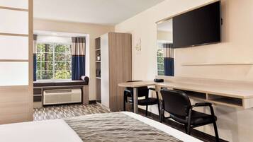 A hotel room with a bed, a desk with two chairs, a TV mounted on the wall, and a window with curtains.