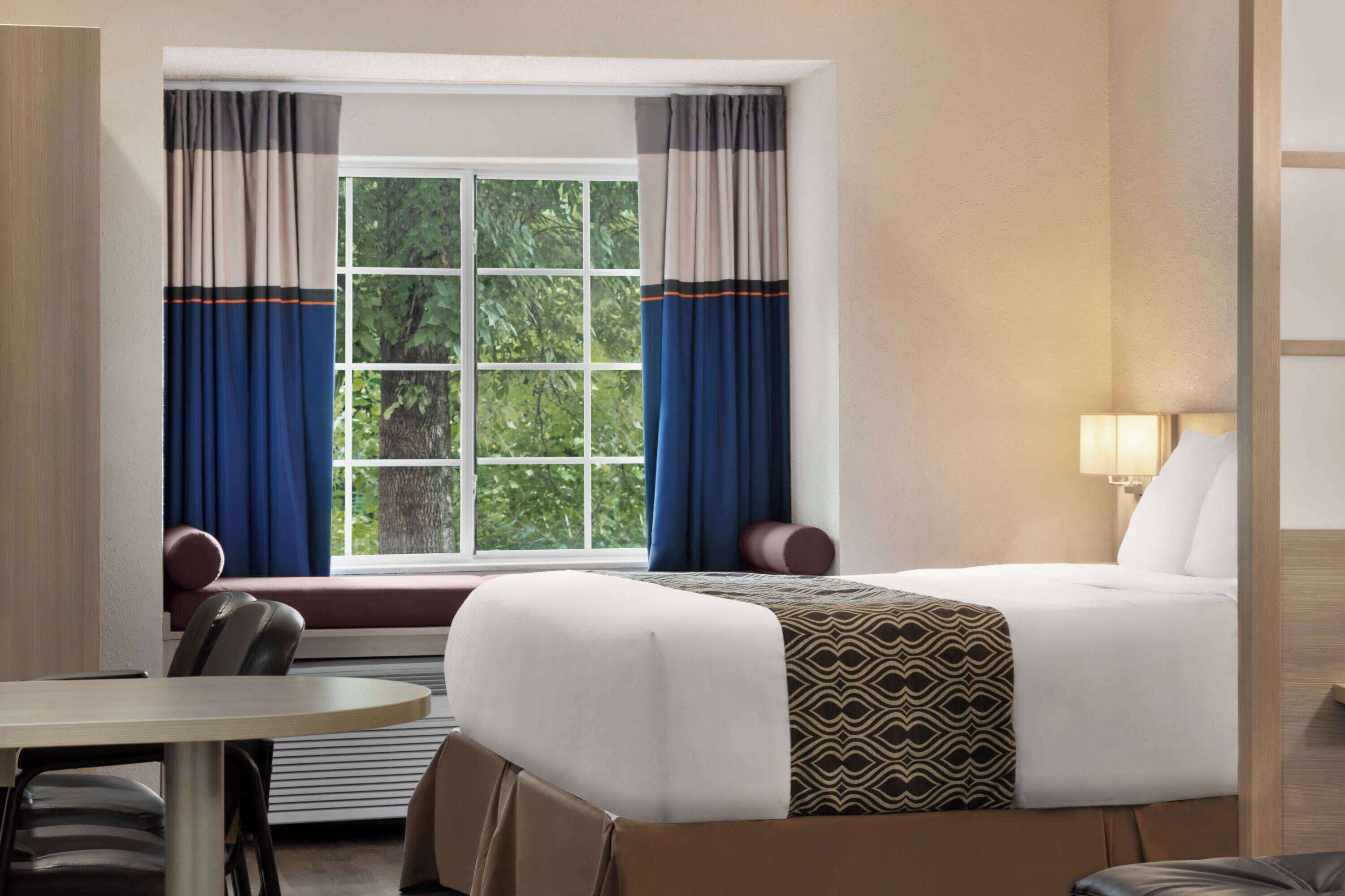 Habitación de hotel con cama, mesita de noche, una silla, ventana con cortinas y vistas a árboles.