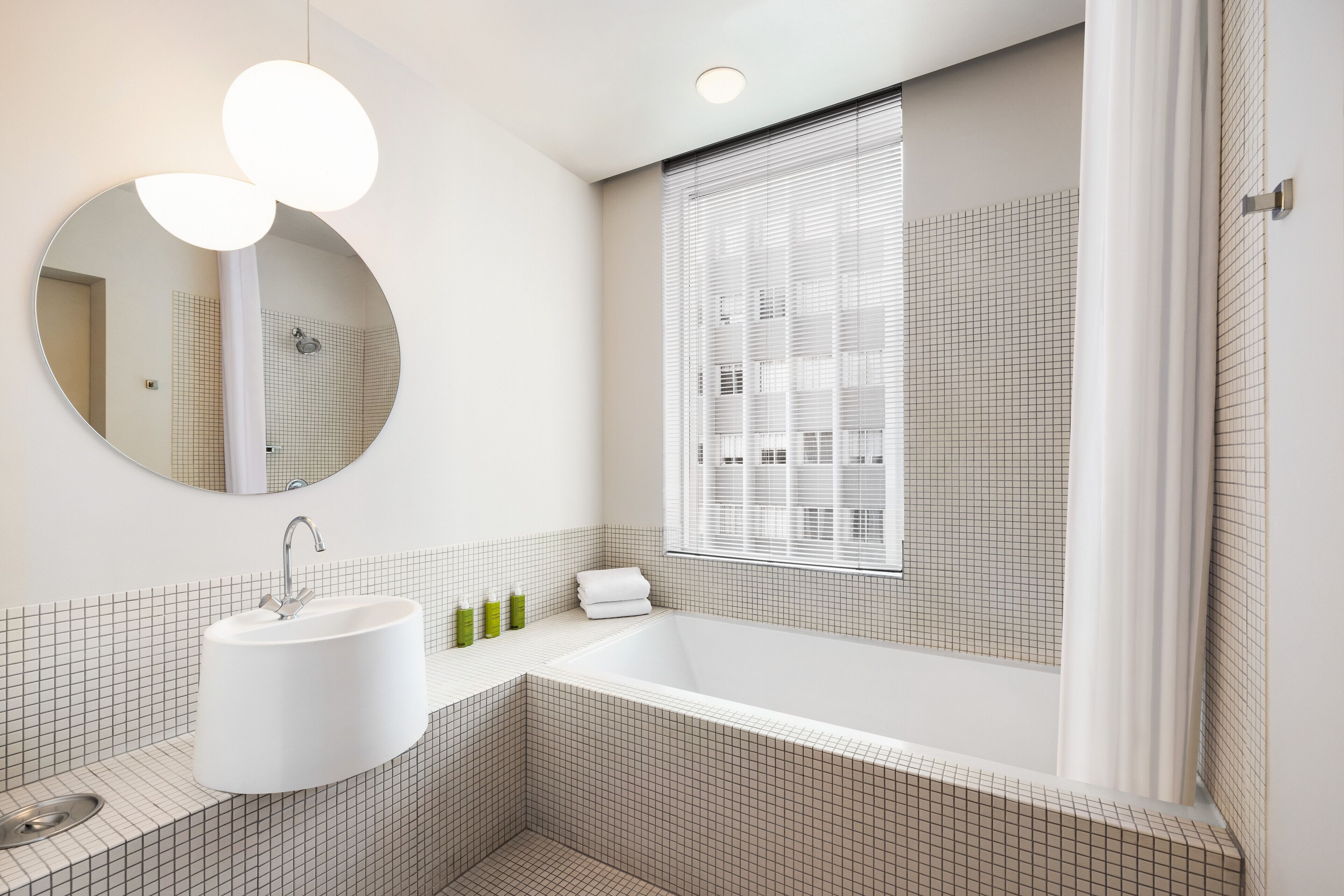 Een moderne badkamer met een vrijstaand bad, een ronde spiegel en een raam met zonwering.