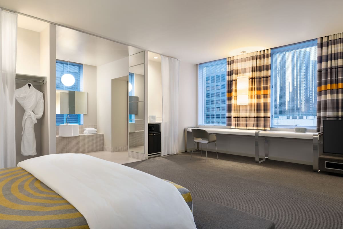 A modern hotel room with a large bed, a desk, a chair, a bathroom with a mirror, and a window with a city view.