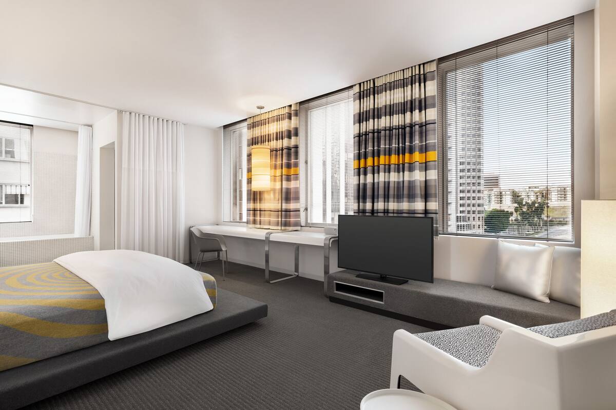 A modern hotel room with a bed, a desk, a TV, and large windows with blinds.