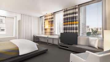 A modern hotel room with a bed, a desk, a TV, and large windows with blinds.