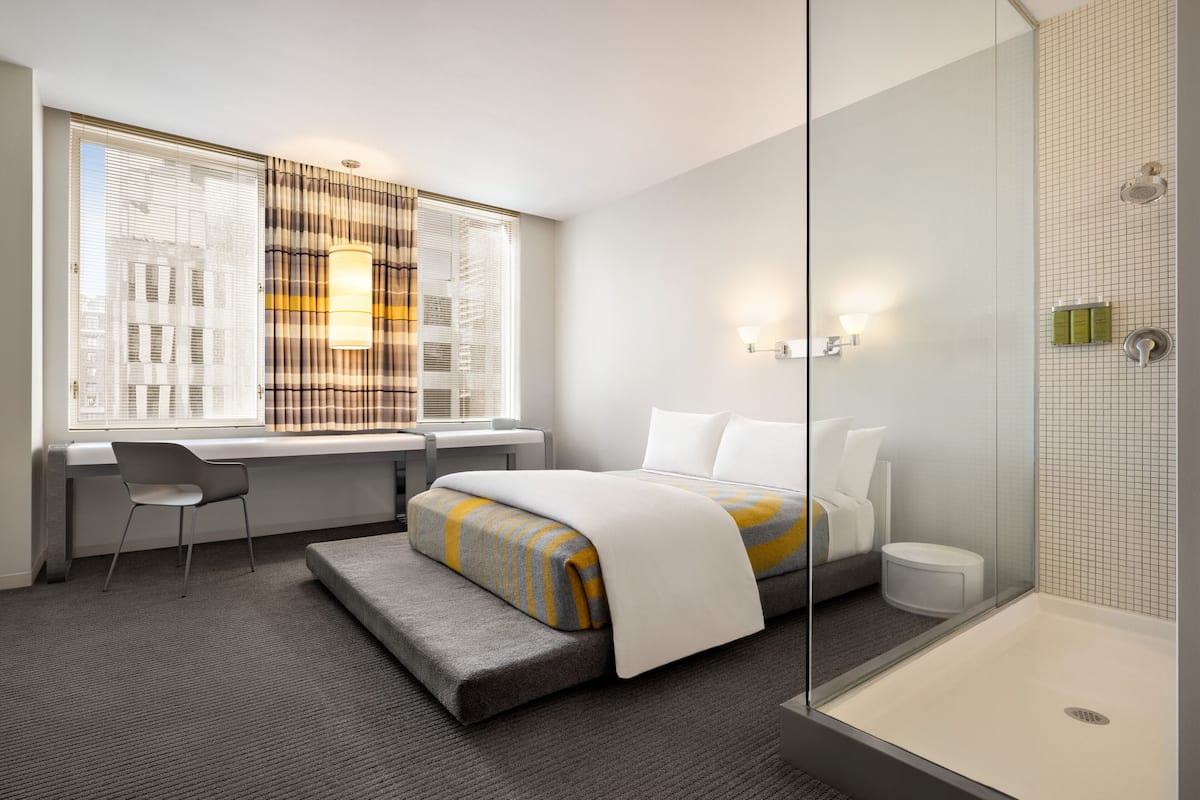 A modern hotel room with a bed, a desk, a chair, a shower, and a window with blinds.