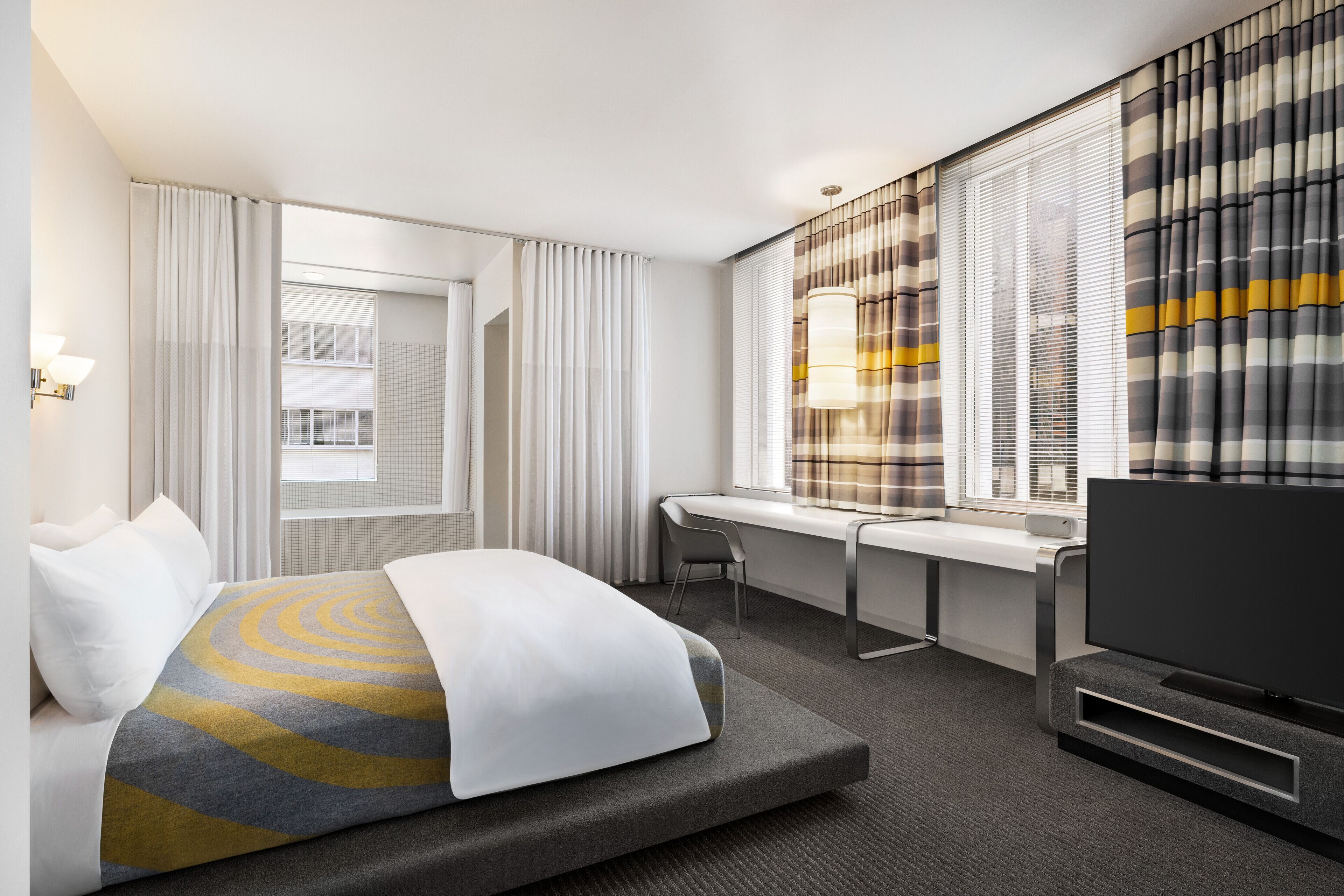 Een moderne hotelkamer met een bed, een bureau, een televisietoestel en grote ramen voorzien van gordijnen.