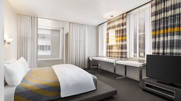Een moderne hotelkamer met een bed, een bureau, een televisietoestel en grote ramen voorzien van gordijnen.