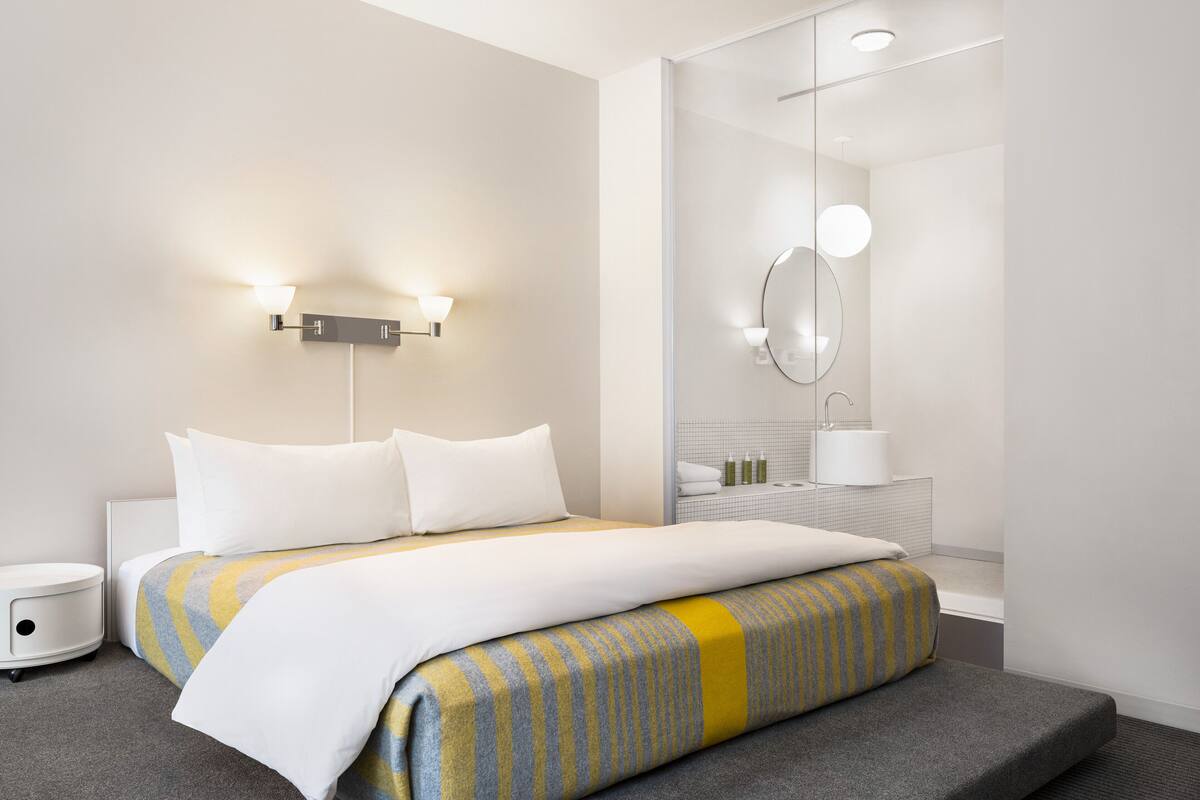 A modern hotel room with a neatly made bed, a bedside table, a wall-mounted light fixture, and a mirror above the sink.