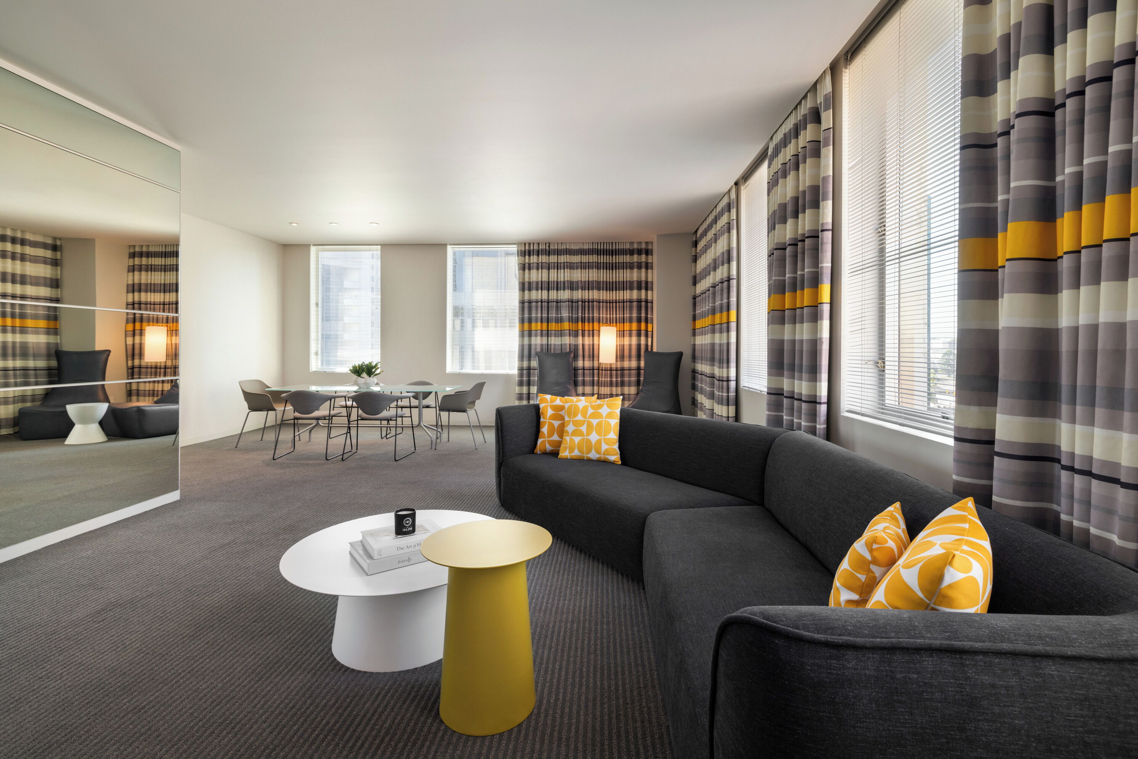 Un salon moderne avec un canapé gris, des coussins aux motifs jaunes et orange, une table basse blanche et un coin repas avec une table et des chaises en verre.