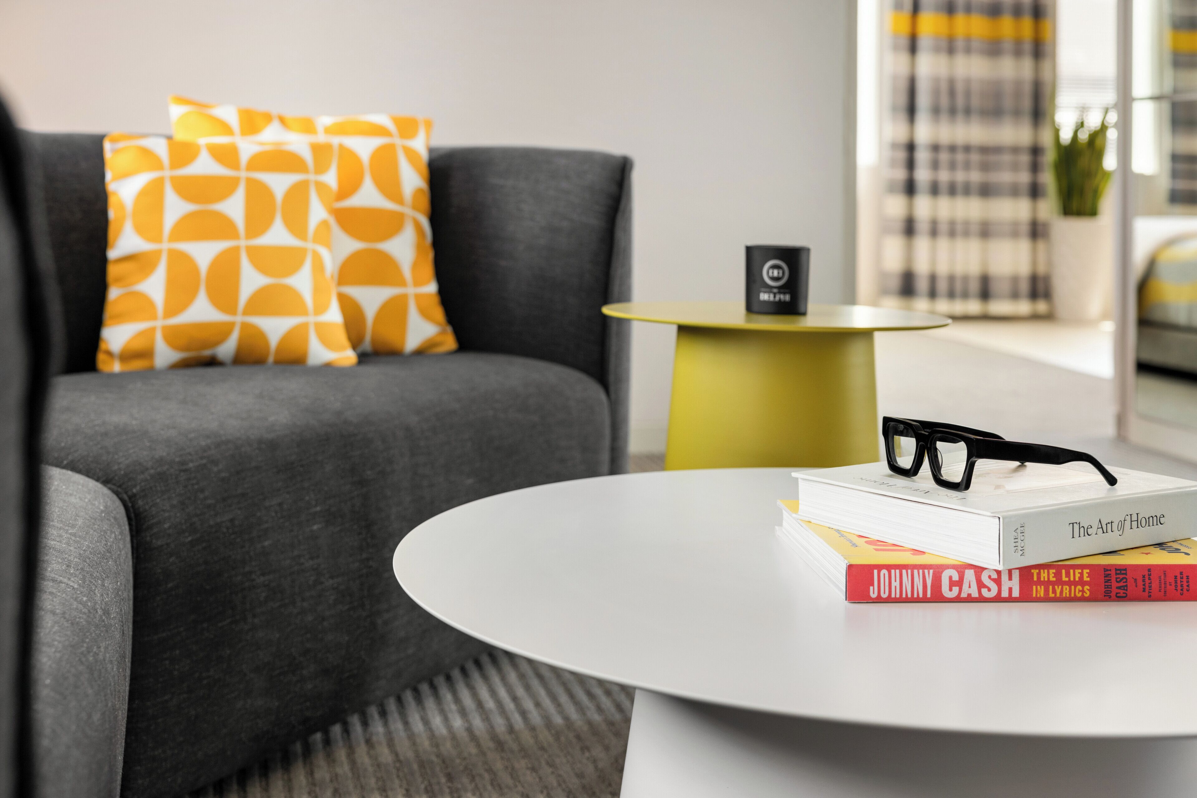 Un salon moderne avec un canapé gris, des coussins à motifs jaunes et blancs, une table basse blanche et des livres, dont « L’art de vivre » et une biographie de Johnny Cash.