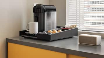 Een koffieapparaat, een witte mok, een dienblad met koffiepads, en een speaker op een aanrecht.