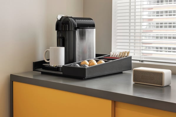 A coffee machine, a white mug, a tray with coffee pods, and a speaker on a counter. - The Delphi Hotel (Los Angeles)