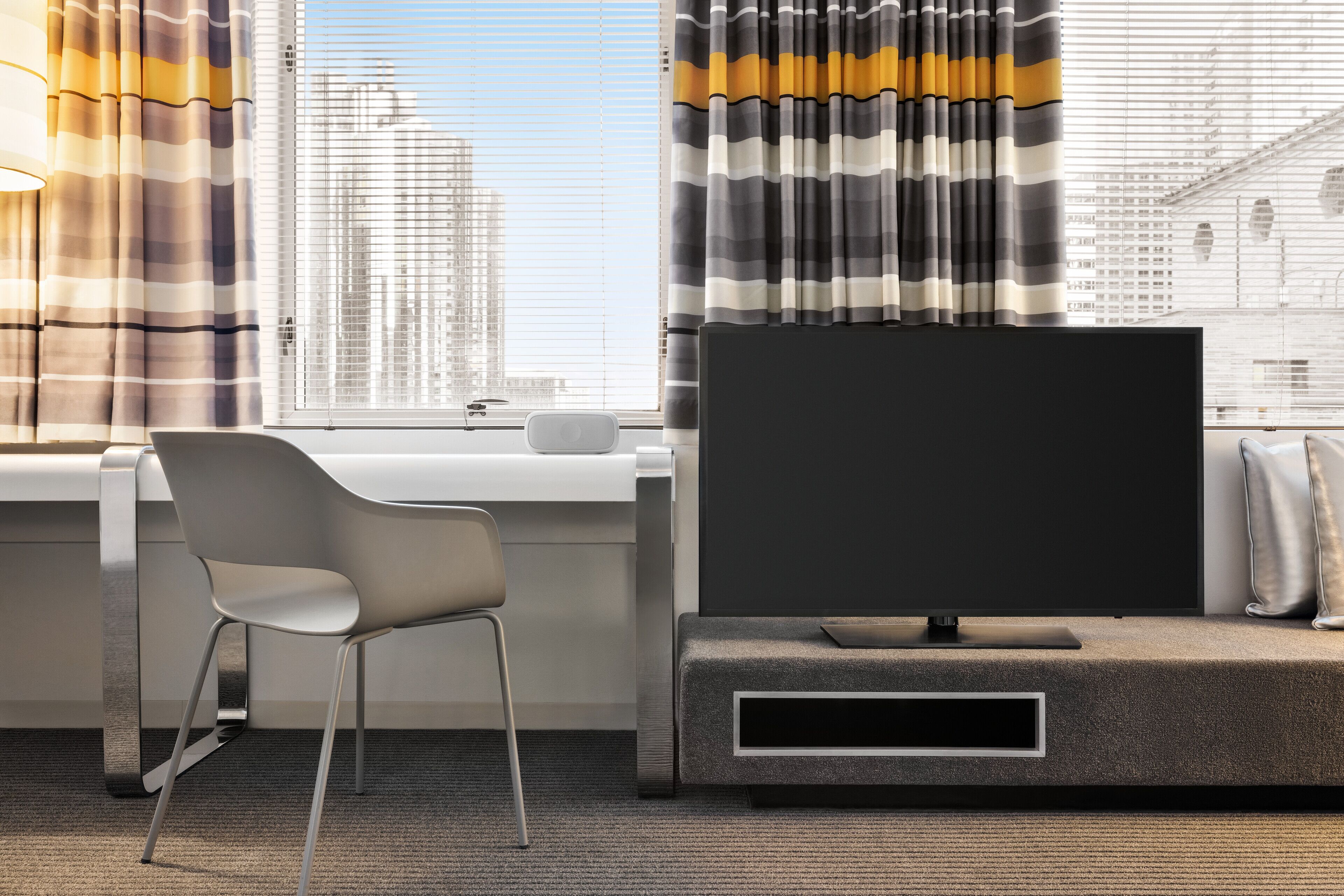 Habitación de hotel moderna con un gran ventanal, televisor de pantalla plana, sofá gris y escritorio blanco.