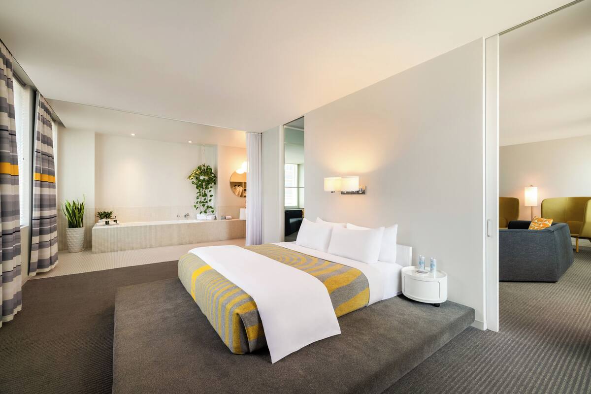 A modern hotel room with a large bed, a sitting area with a sofa, a small table, and a bathroom visible in the background.