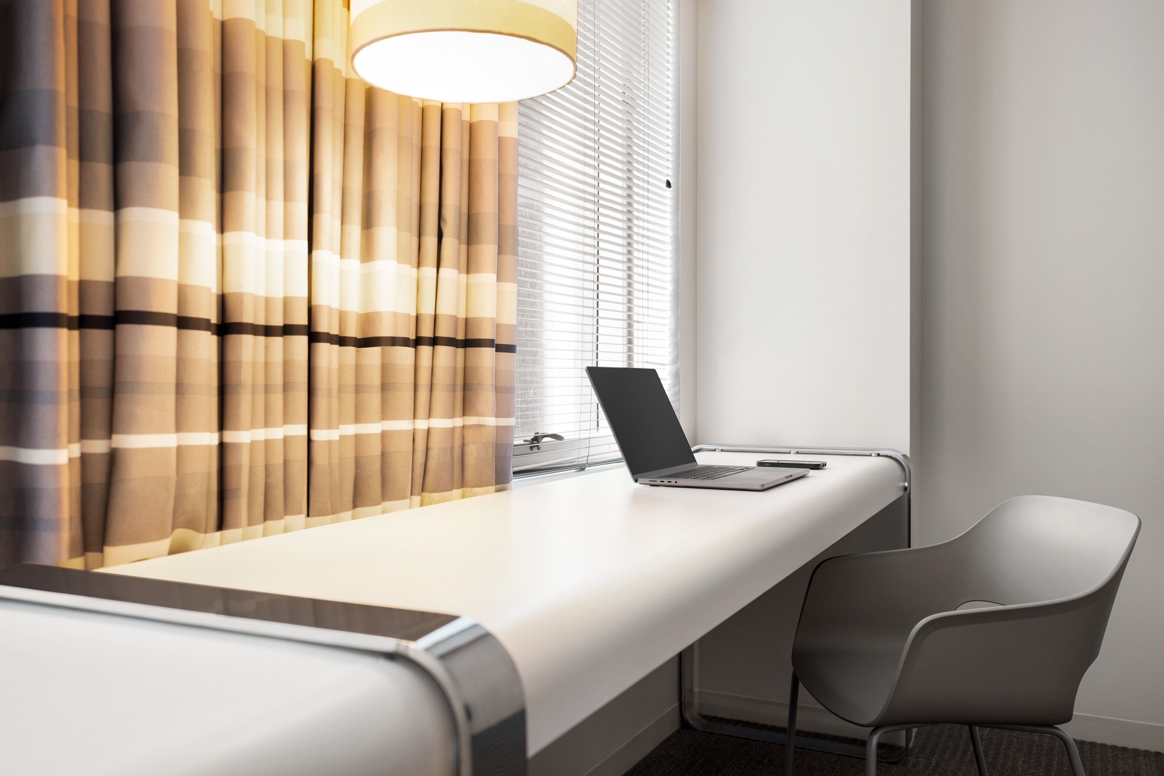 Une chambre d’hôtel moderne avec un grand bureau, un ordinateur portable, une chaise et une fenêtre équipée de stores.