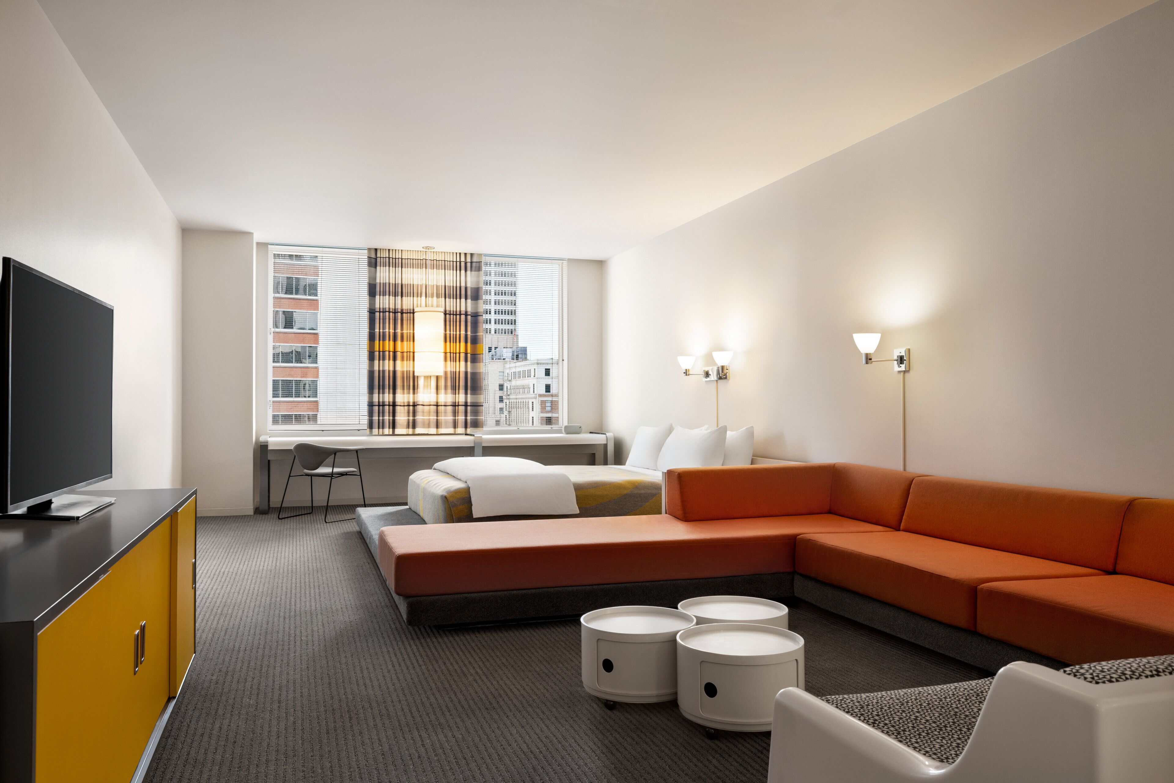 A modern hotel room with a sectional sofa, a flat-screen TV, a desk, and a large window with city views.