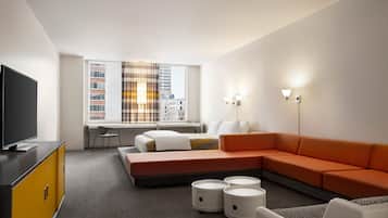 A modern hotel room with a sectional sofa, a flat-screen TV, a desk, and a large window with city views.