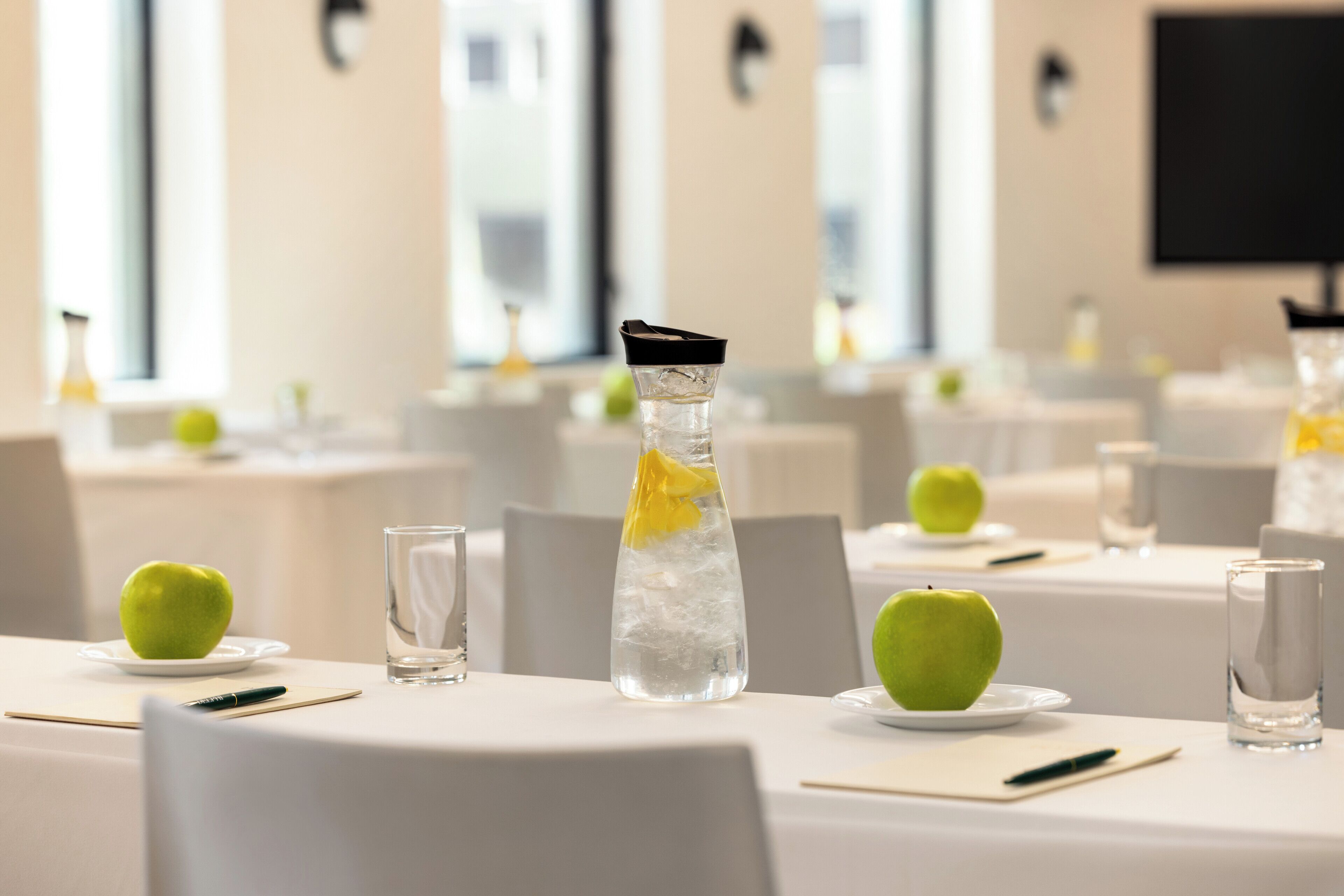 A conference room with tables set for a meeting, each with a glass of water, a plate with an apple, and a notepad.