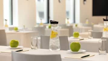 Un salón de conferencias con mesas dispuestas para una reunión, cada una con un vaso de agua, un plato con una manzana y un bloc de notas.