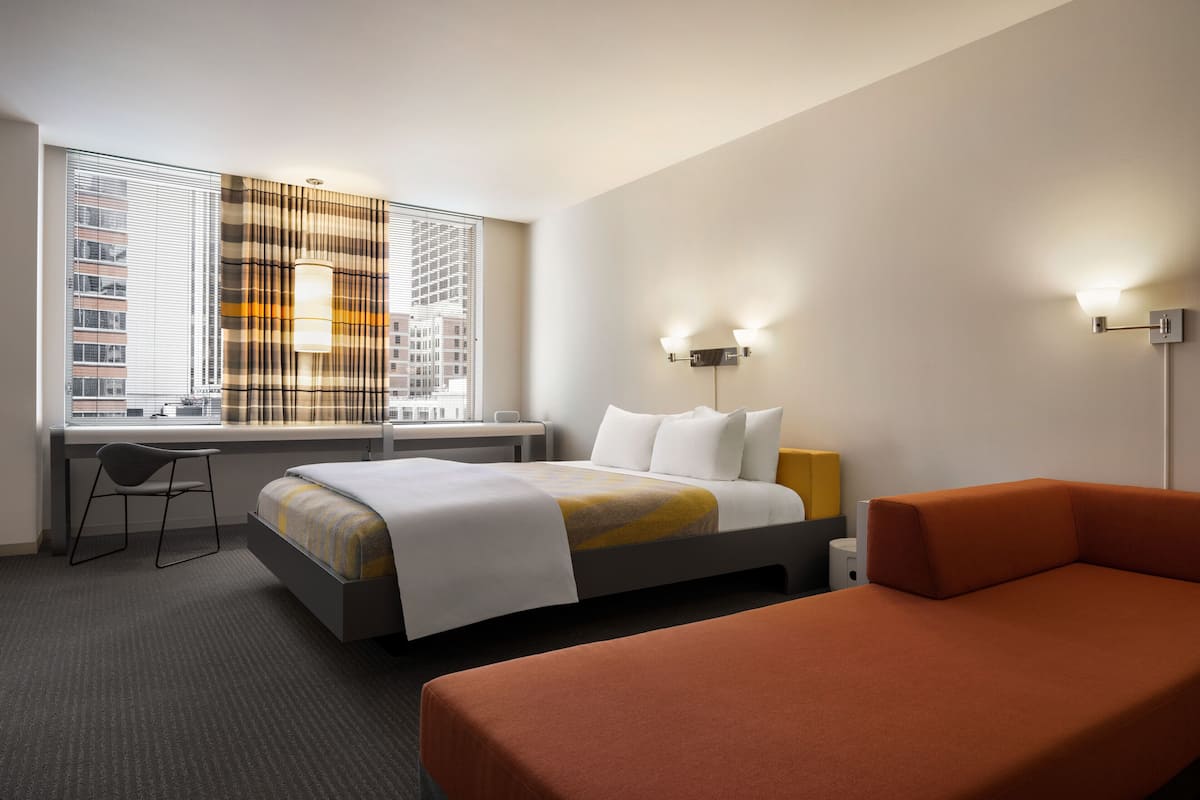 A hotel room with a bed, a desk, a chair, a sofa, and a window with blinds.