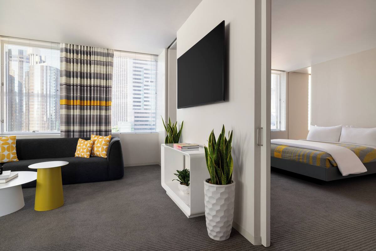 A modern hotel room with a sofa, a small white table, a potted plant, a bed with a striped bedspread, and a large window with blinds.