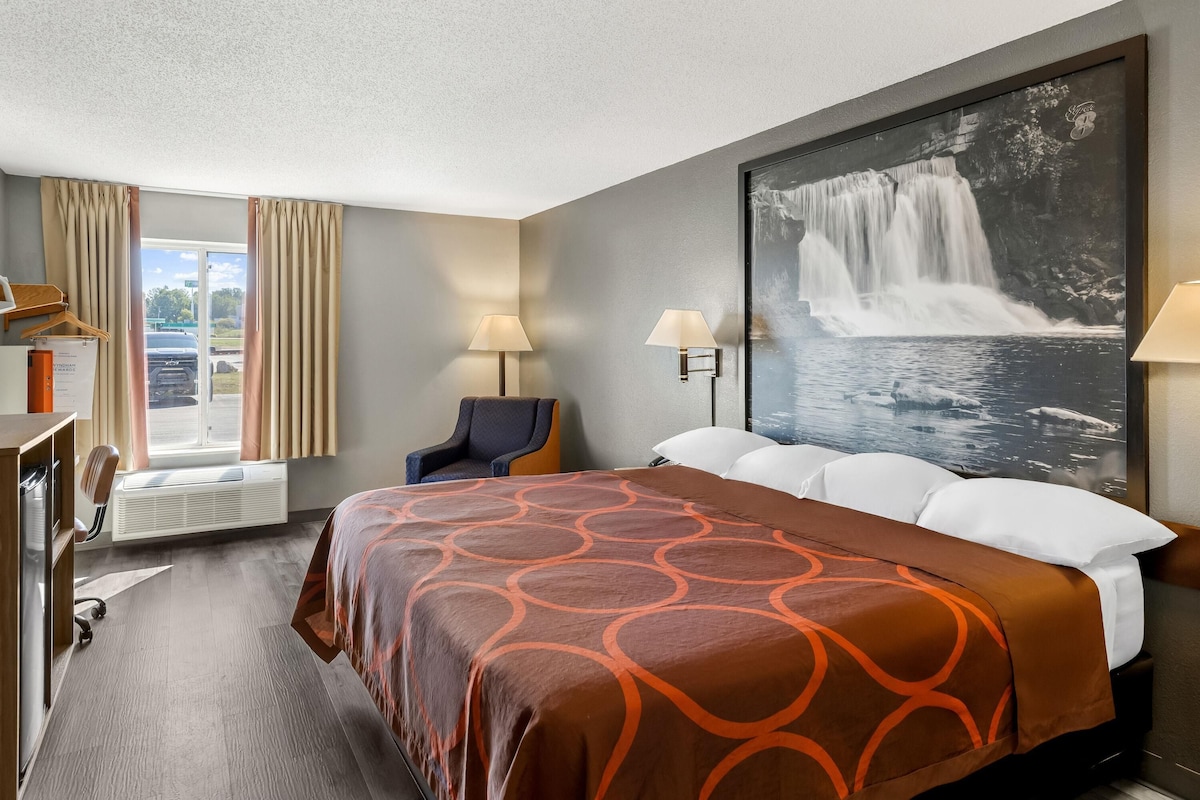 A hotel room with a large bed, two bedside lamps, a chair, a desk with a lamp, a large window with curtains, and a wall-mounted TV.