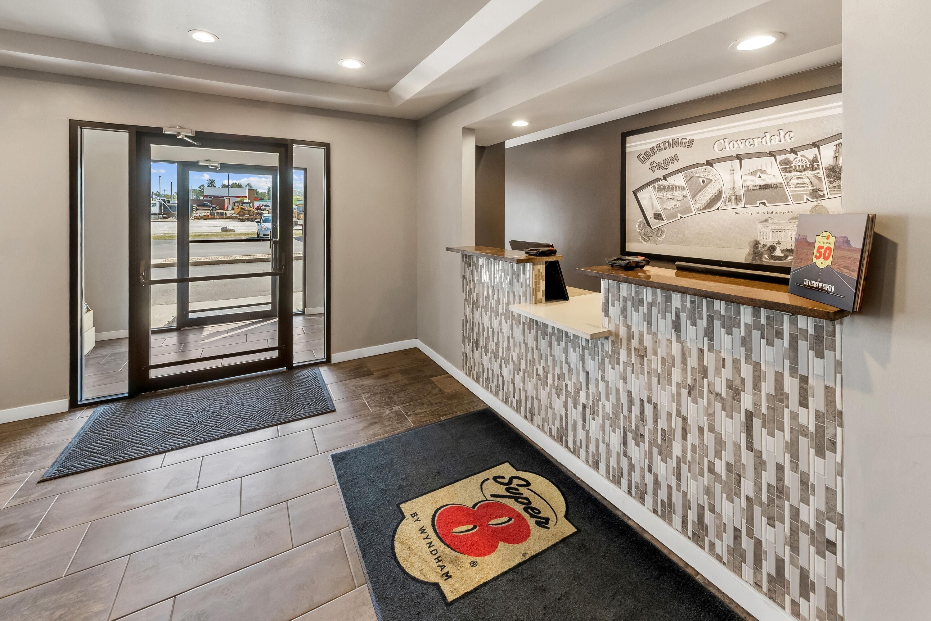 A hotel reception area with a glass door entrance, a reception desk, a wall-mounted display, and a doormat.