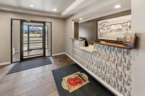 Zona de recepción de hotel con entrada por puerta de vidrio, mostrador de recepción, panel de pared y felpudo.
