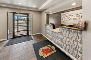 Zona de recepción de hotel con entrada por puerta de vidrio, mostrador de recepción, panel de pared y felpudo.