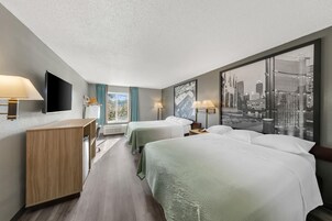 Habitación de hotel con dos camas, un escritorio, un televisor de pantalla plana y un ventanal con vista a la ciudad.