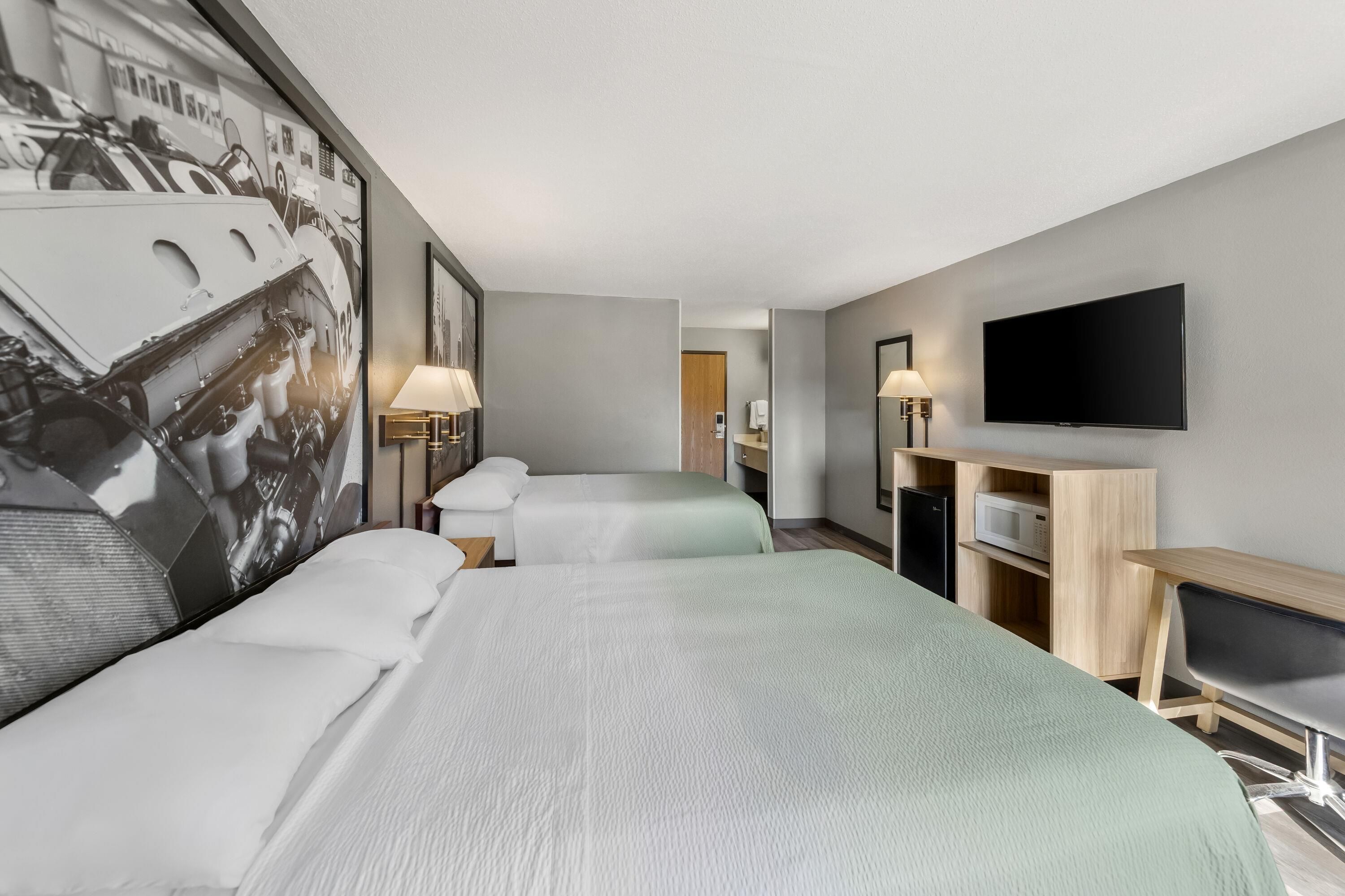 A hotel room with two beds, a television, a desk with a computer, and a wall-mounted TV.