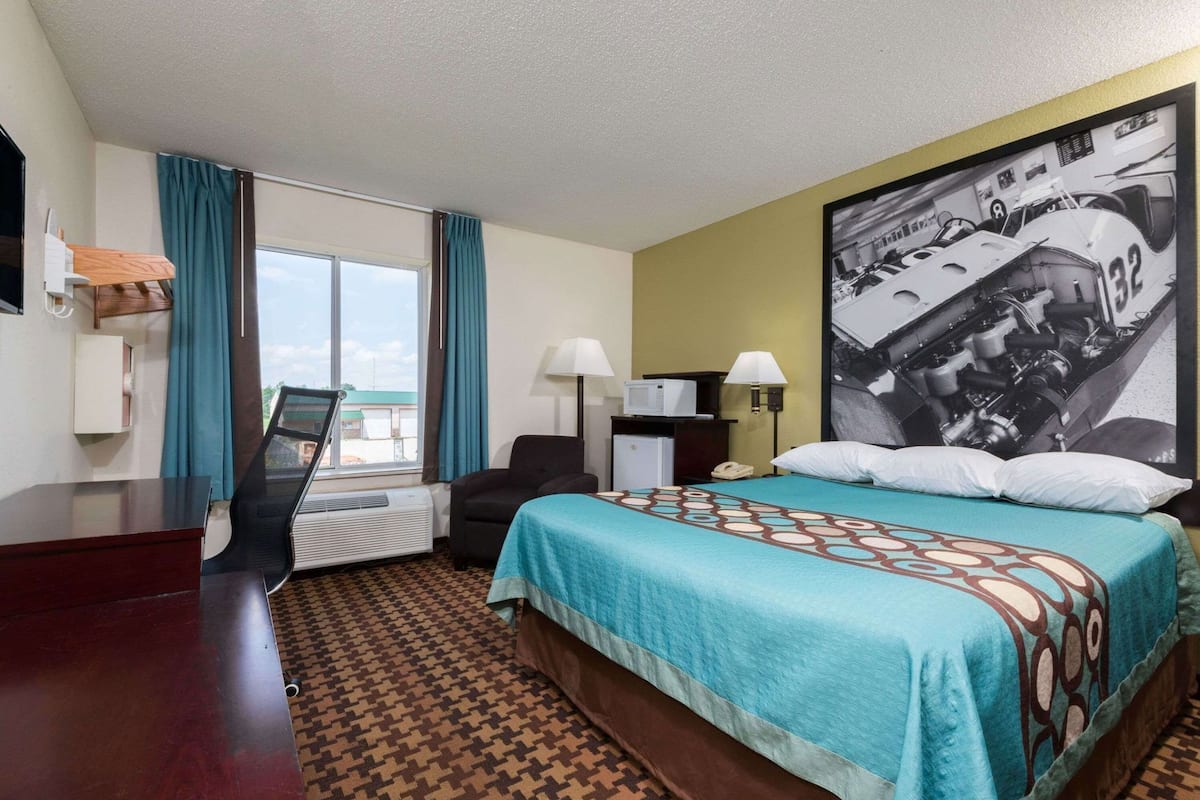 A hotel room with a bed, a desk, a chair, a window with curtains, a wall-mounted TV, a lamp, and a large poster on the wall.