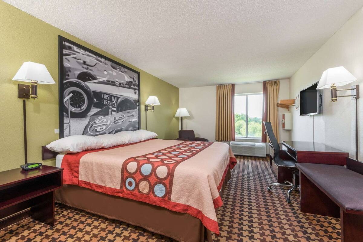 A hotel room with a bed, two bedside lamps, a desk, a chair, a TV, and a large poster on the wall.