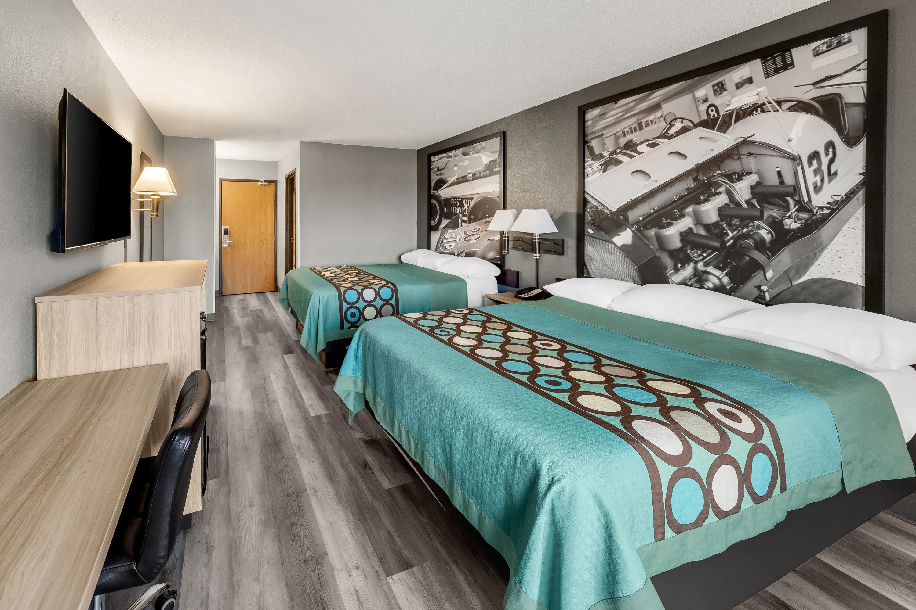 A hotel room with two beds, a desk, a TV, and a wall with a car and engine image.