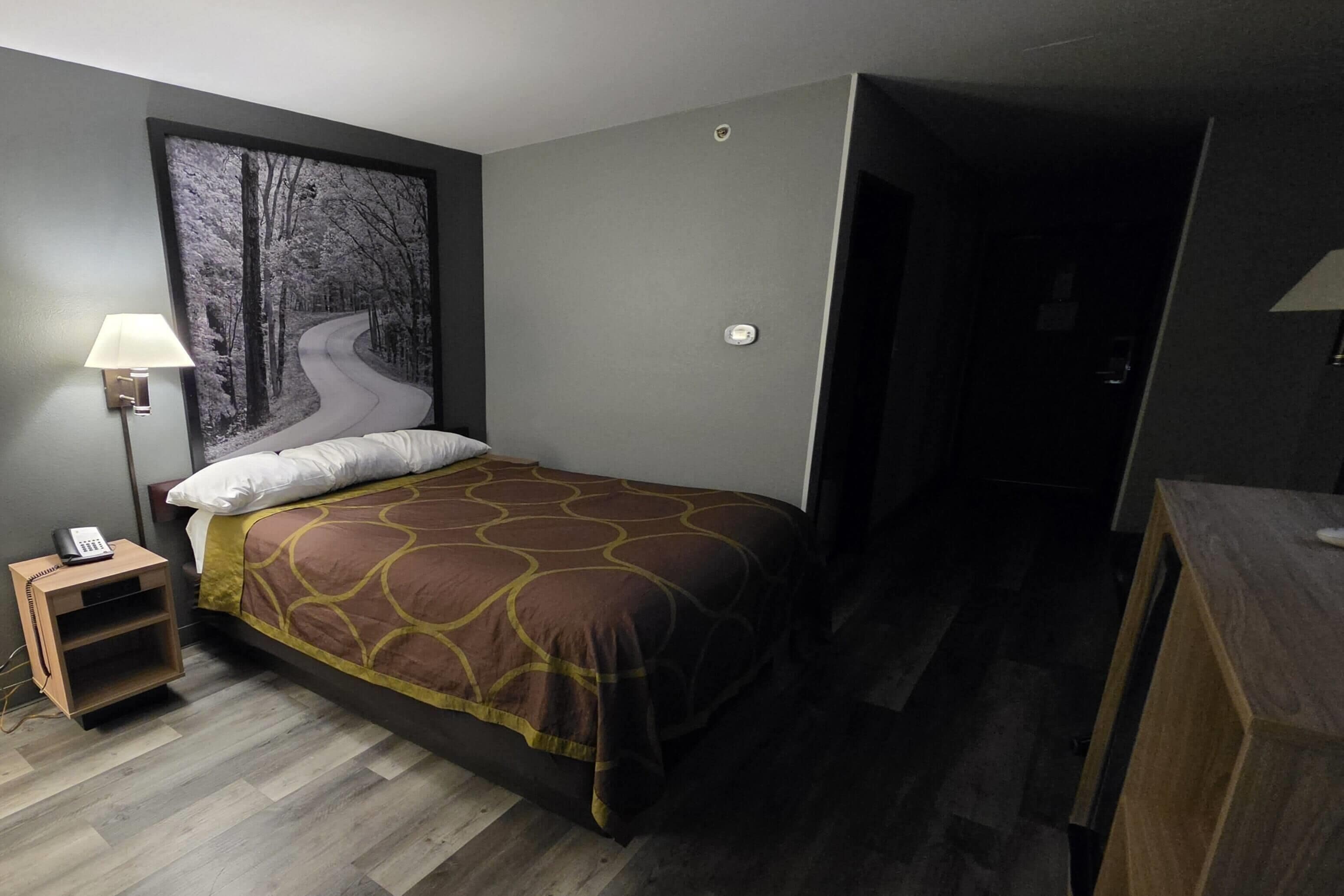 Habitación de hotel con cama, mesita de noche, lámpara y un cuadro de un camino en el bosque.
