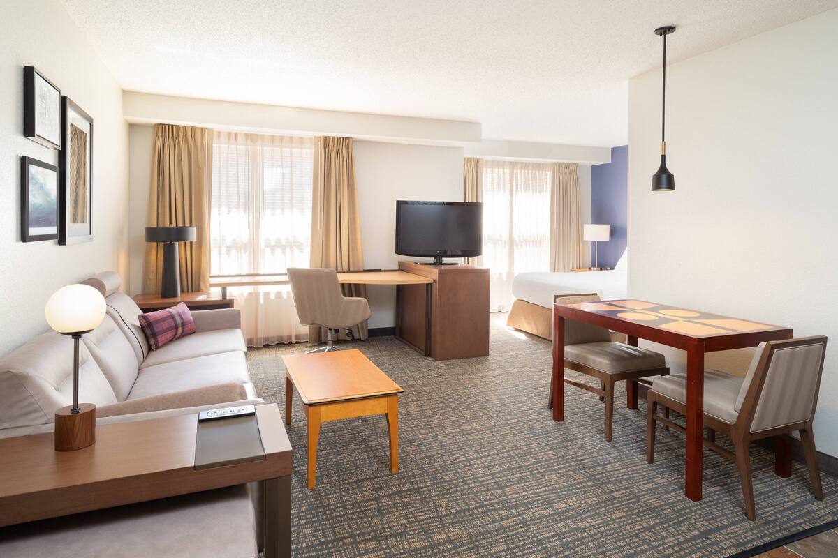 A hotel room with a sofa, a desk with a computer, a TV, a dining table with chairs, and a lamp.