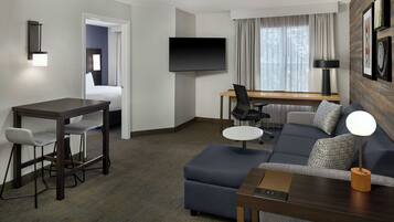 A hotel room with a flat-screen TV, a dining table with chairs, a sofa, and a small round table with a lamp.