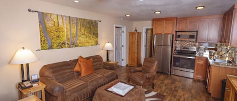 A living room with a sofa, armchair, and coffee table, a kitchen with modern appliances, and a ceiling fan.
