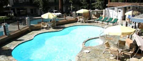 Piscina con sillas de descanso y sombrillas, rodeada de baldosas de piedra y árboles.