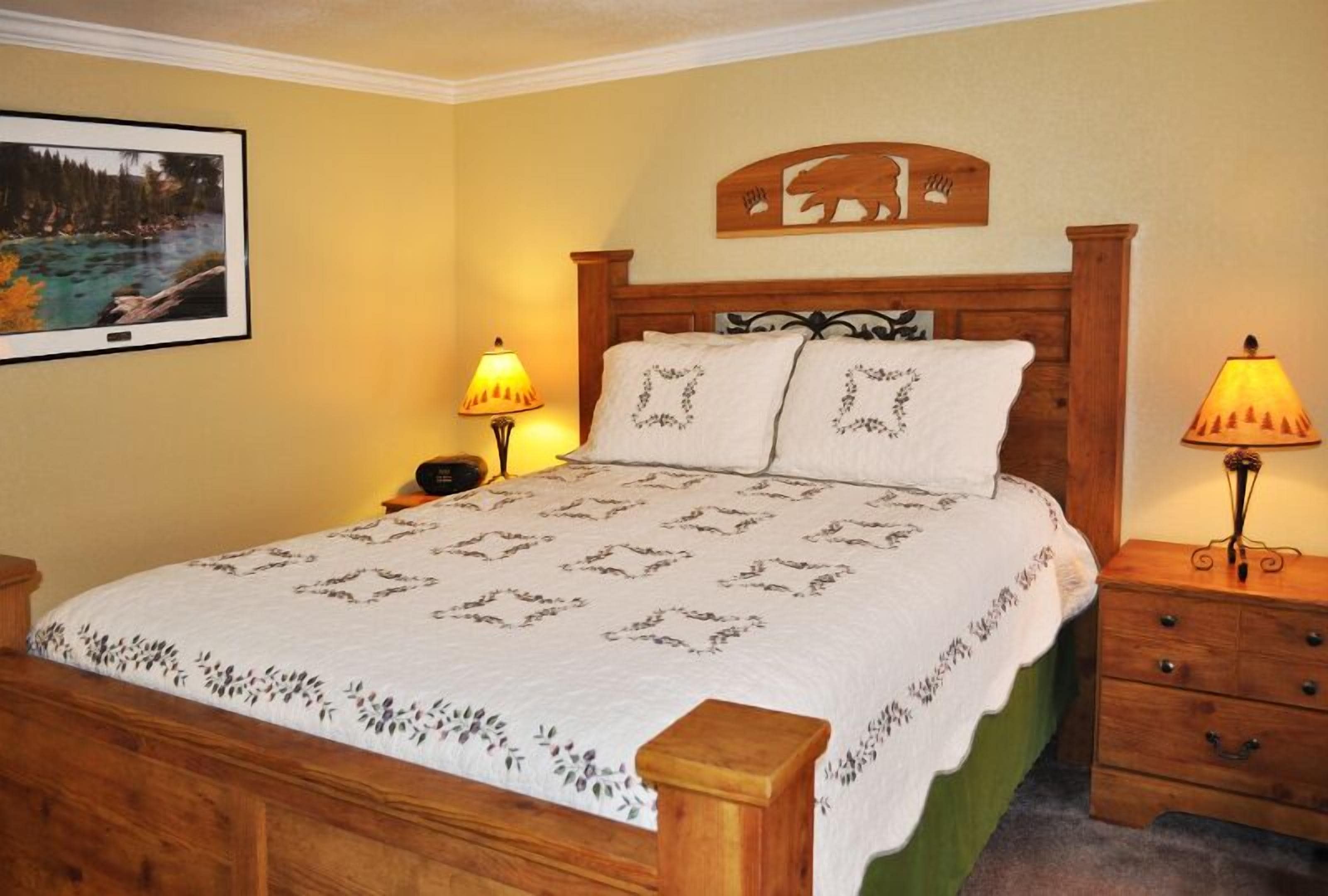 A bedroom with a wooden bed, two bedside lamps, a framed picture on the wall, and a wooden headboard with a bear design.
