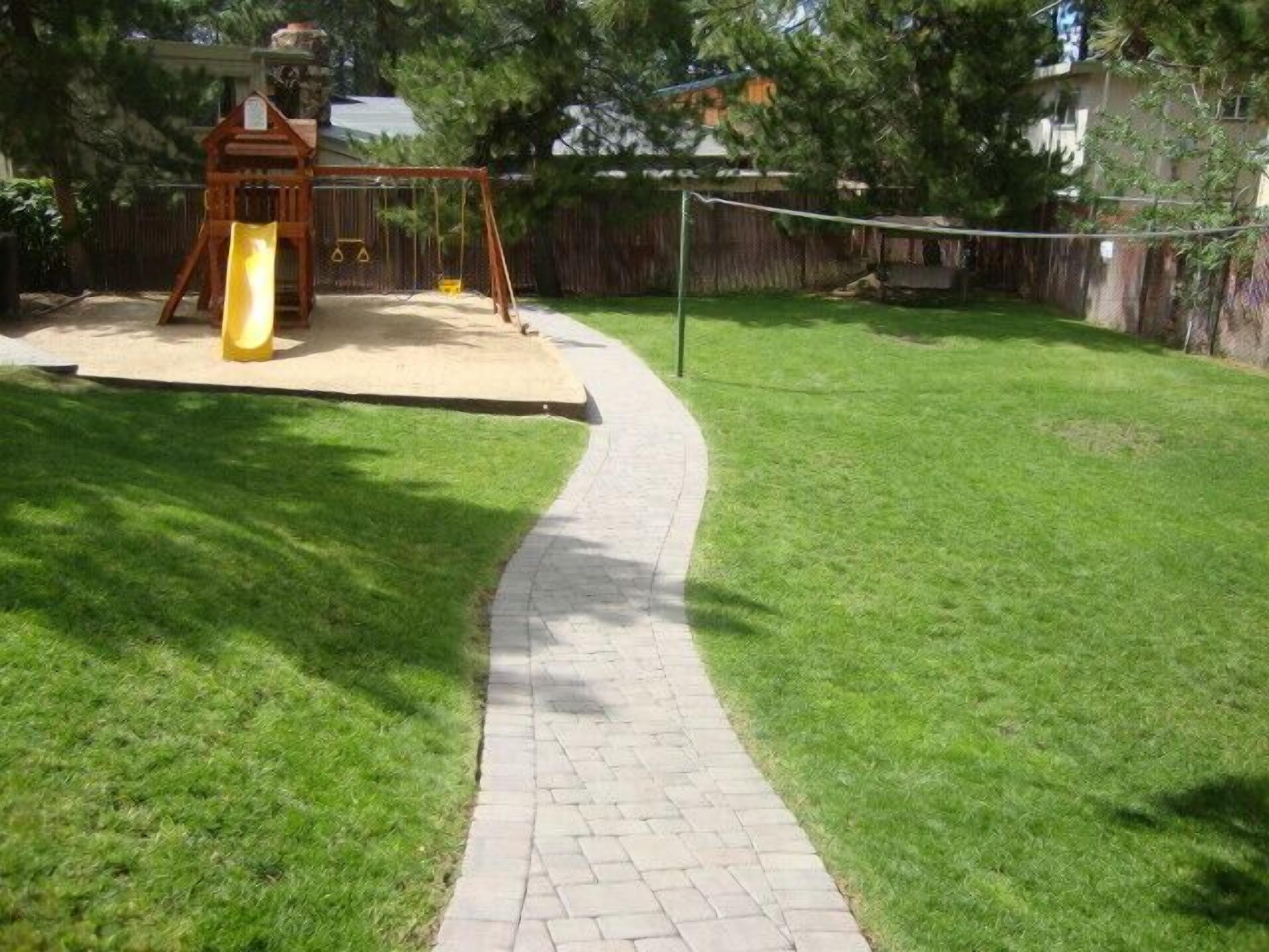 A backyard with a paved path, a wooden swing set, and a fenced area.