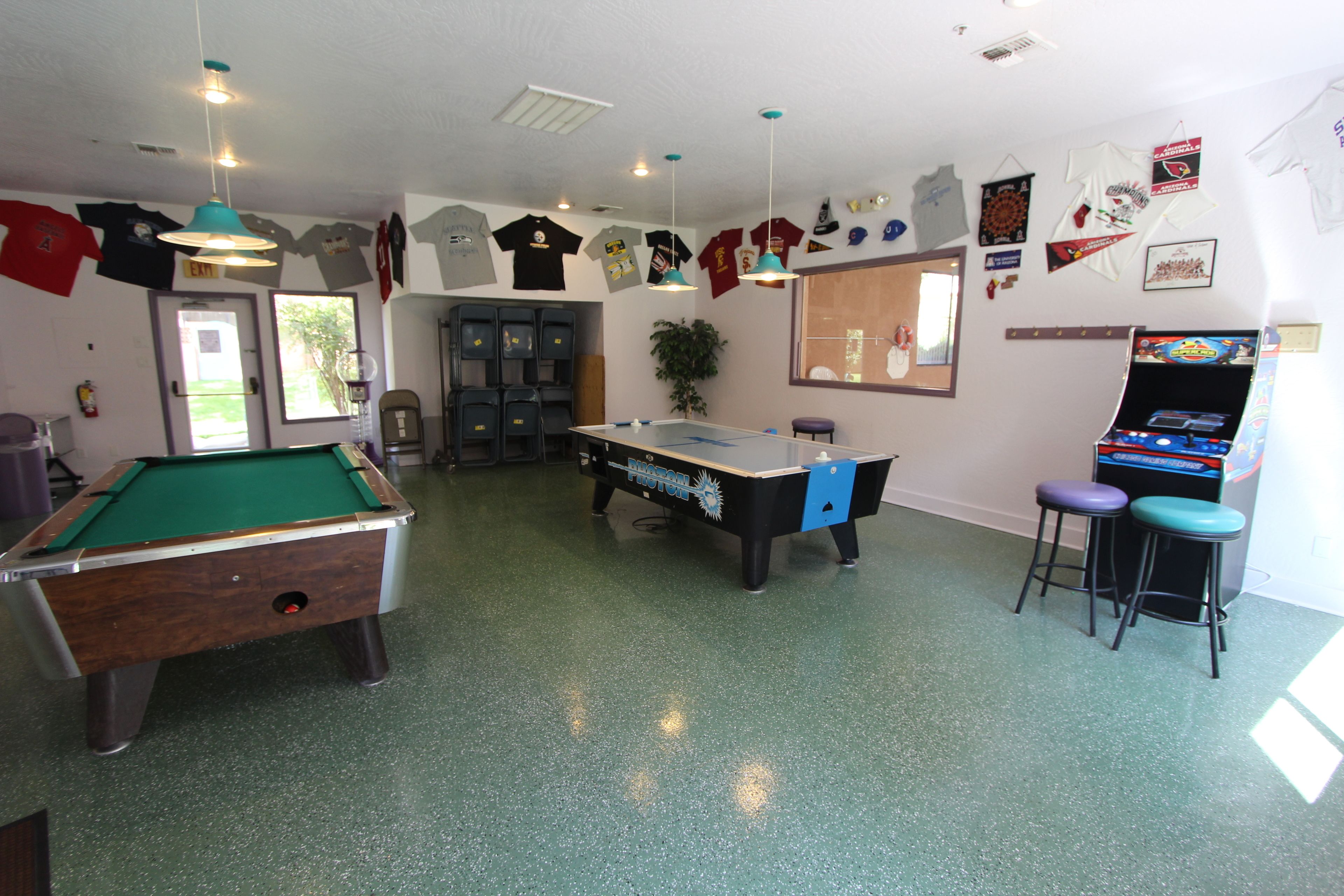 A pool table, a foosball table, and a bar area with stools in a room with green flooring and various sports memorabilia on the walls.