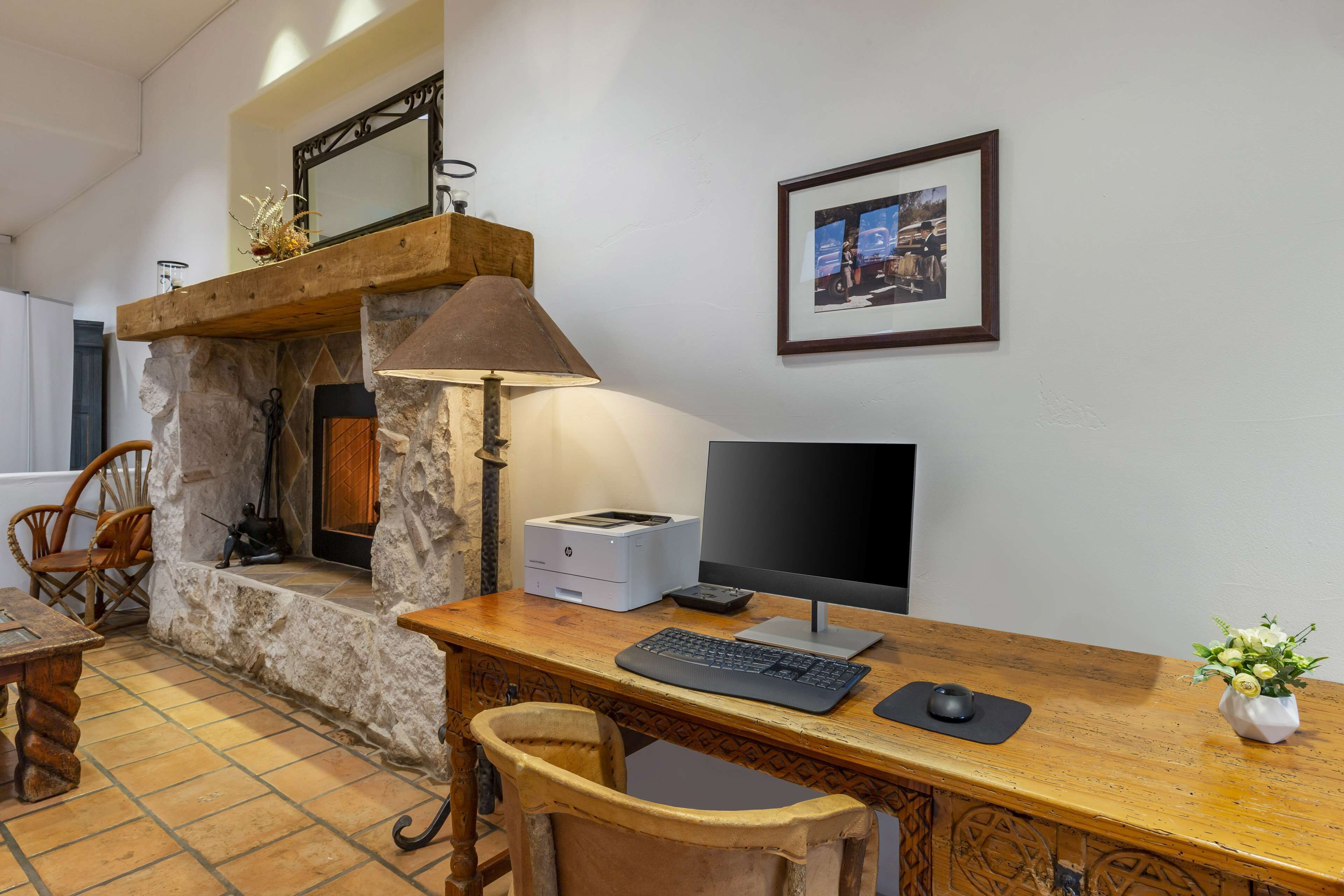 A rustic room with a stone fireplace, a wooden desk with a computer, a lamp, and a framed picture on the wall.