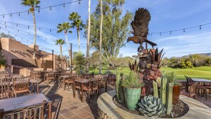 An outdoor patio with wooden tables and chairs, surrounded by cacti and a large bird sculpture.