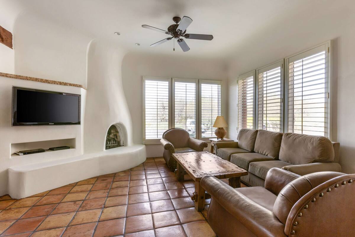 A living room with a flat-screen TV, a ceiling fan, a fireplace, a sofa, a coffee table, and large windows with blinds.
