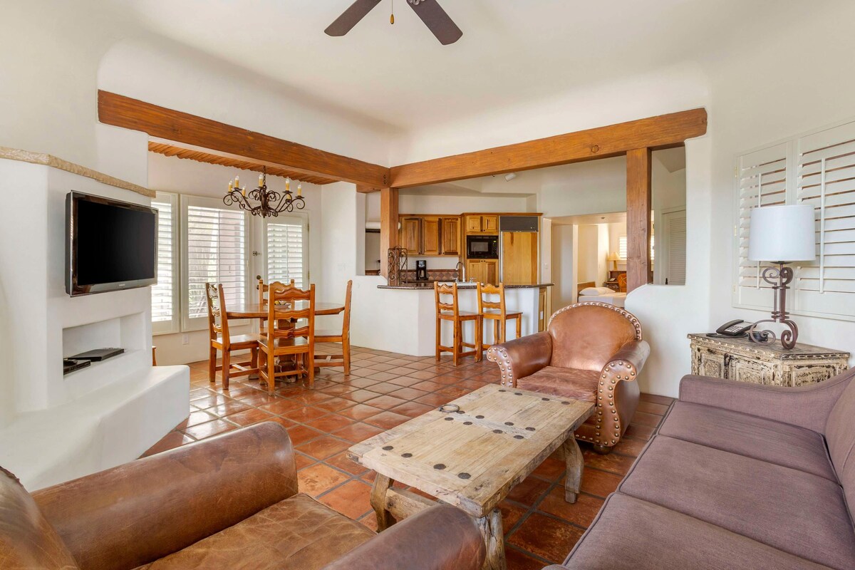 A living room with a television, wooden furniture, and a kitchen in the background.