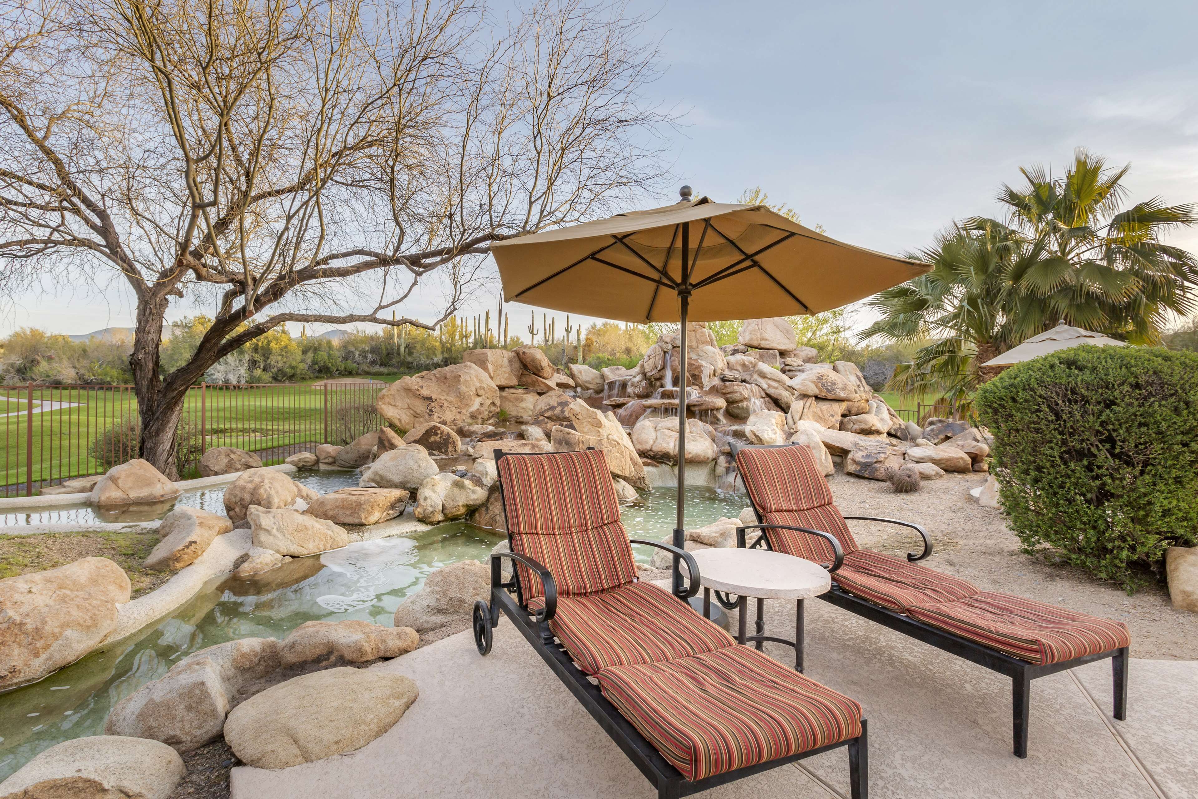A patio with two lounge chairs and a table, a large umbrella, and a small water feature surrounded by rocks.