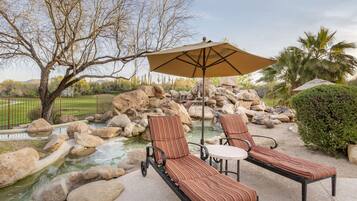 A patio with two lounge chairs and a table, a large umbrella, and a small water feature surrounded by rocks.